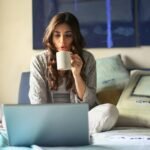 A woman enjoying coffee while working from home in a cozy bedroom setting with a laptop.