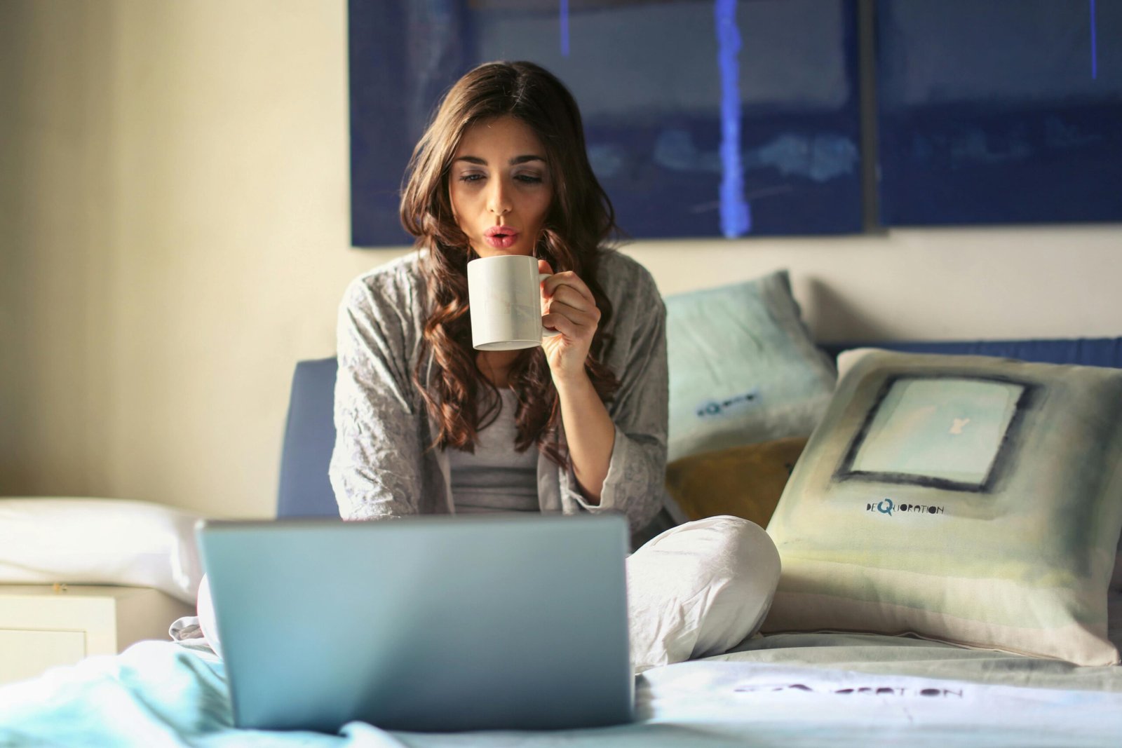 A woman enjoying coffee while working from home in a cozy bedroom setting with a laptop.